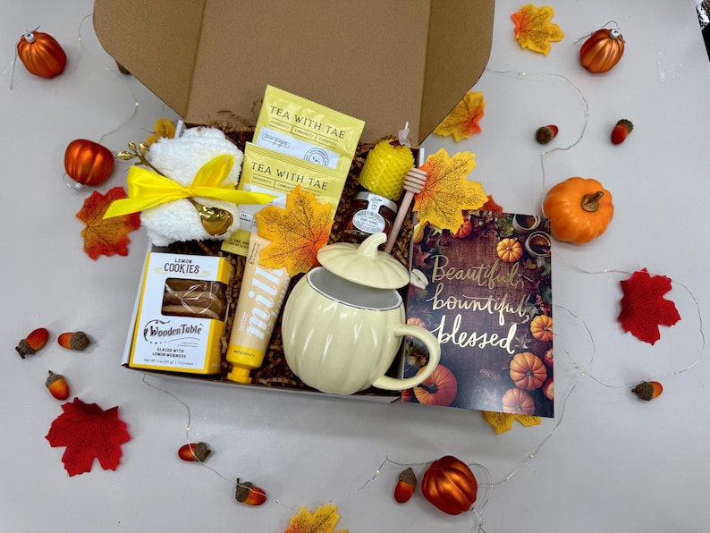 May include: A gift box filled with fall-themed items. Includes a pumpkin-shaped mug, cookies, tea, a candle, and a card with the words "Beautiful, bountiful, blessed." Decorative leaves, pumpkins, and acorns surround the box.