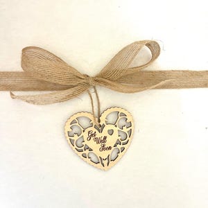 Pode incluir: Um ornamento de madeira em forma de cora&ccedil;&atilde;o com um design delicado de corte. O ornamento &eacute; amarrado com um la&ccedil;o de corda marrom e tem as palavras "Get Well Soon" escritas nele.