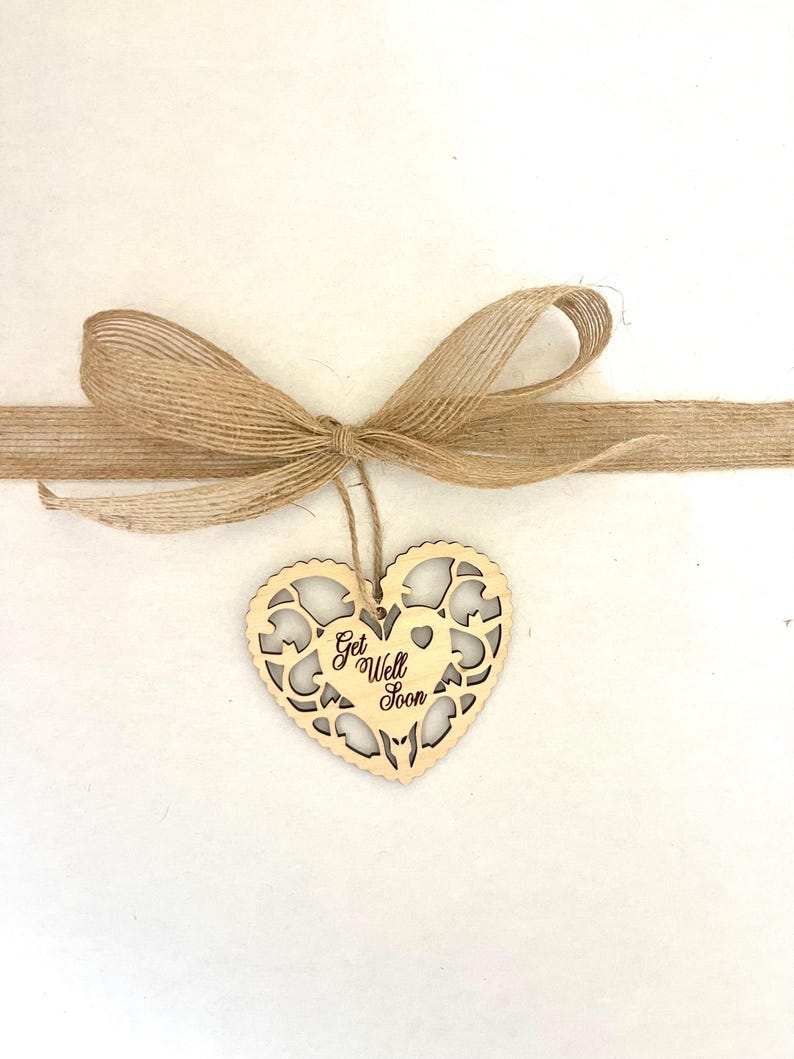 Pode incluir: Um ornamento de madeira em forma de cora&ccedil;&atilde;o com um design delicado de corte. O ornamento &eacute; amarrado com um la&ccedil;o de corda marrom e tem as palavras "Get Well Soon" escritas nele.