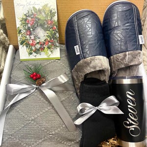 Pu&ograve; includere: Una scatola regalo piena di una coperta di maglia grigia, un paio di pantofole blu con fodera in pelliccia sintetica, calzini neri e un bicchiere in acciaio inossidabile con il nome "Steven" inciso. La scatola contiene anche un biglietto di Natale con una ghirlanda.