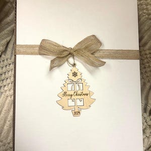 May include: A white card with a brown burlap ribbon and bow. A wooden Christmas tree ornament hangs from the ribbon. The ornament is cut out in the shape of a Christmas tree with a present inside and the words "Merry Christmas!" and "2024" written on it.