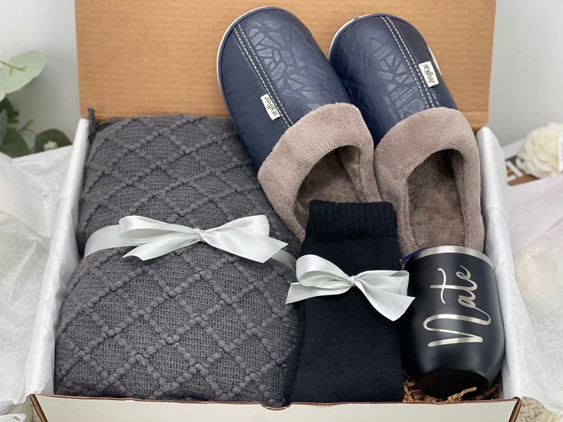 Pode incluir: Uma caixa de presente cheia de itens aconchegantes, incluindo uma manta de malha cinza, um par de pantuflas azul escuro com forro de l&atilde; cinza, um par de meias pretas e uma caneca de viagem preta com a palavra "Nate" gravada.