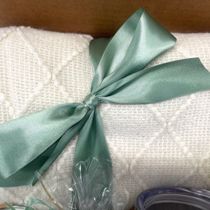May include: A white knitted throw blanket with a diamond pattern is tied with a sage green satin ribbon bow.