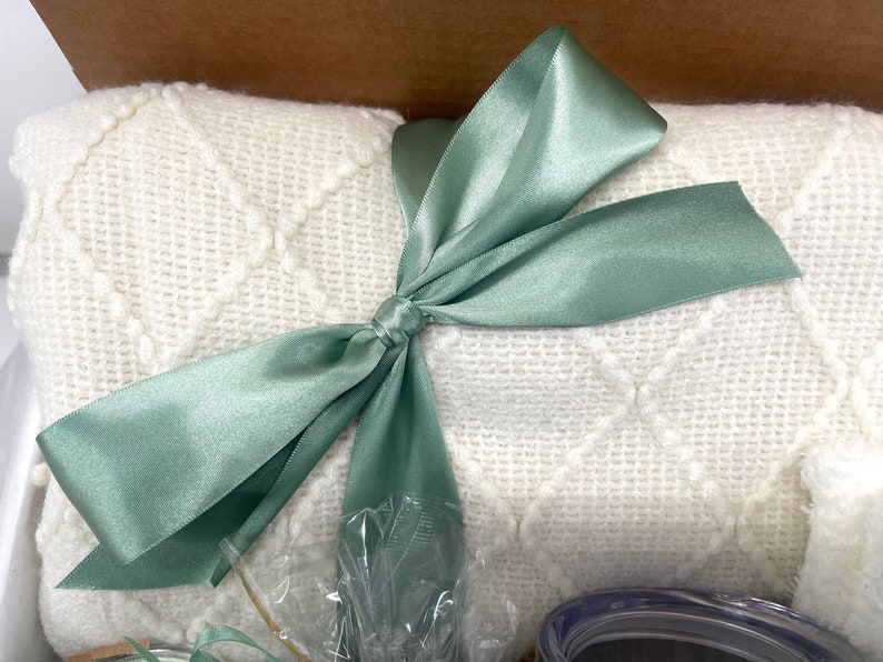 May include: A white knitted throw blanket with a diamond pattern is tied with a sage green satin ribbon bow.