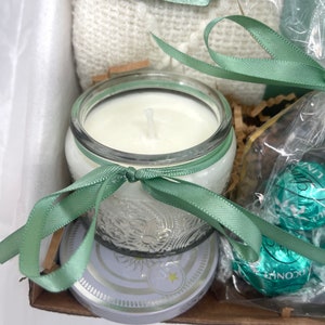 May include: A gift basket with a white blanket, a clear glass candle with a white wax, a green ribbon, and a box of chocolates wrapped in green and silver foil.