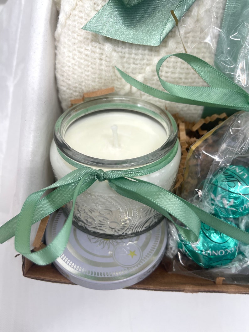 May include: A gift basket with a white blanket, a clear glass candle with a white wax, a green ribbon, and a box of chocolates wrapped in green and silver foil.
