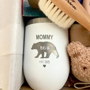 Może przedstawiać: Biały, izolowany kubek z nadrukiem "MOMMY BEAR" i "EST. 2025". Kubek ma sylwetkę niedźwiedzia. Widoczna jest r&oacute;wnież drewniana szczotka dla dziecka z napisem "Baby Anderson".