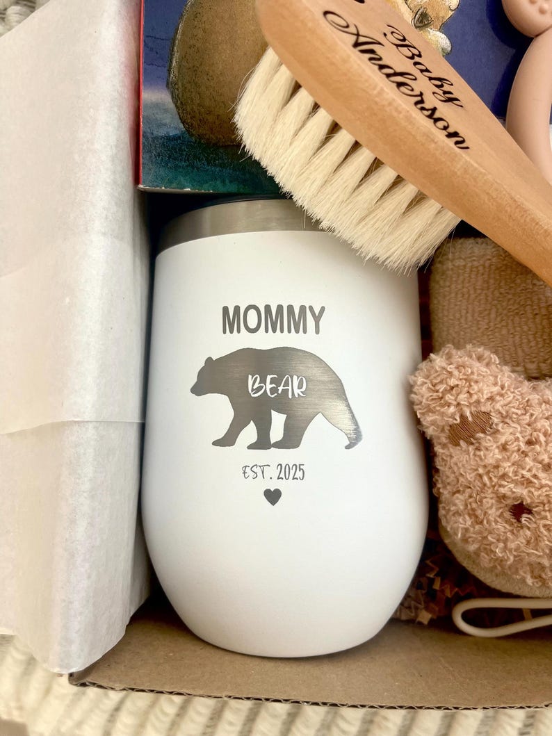 Może przedstawiać: Biały, izolowany kubek z nadrukiem "MOMMY BEAR" i "EST. 2025". Kubek ma sylwetkę niedźwiedzia. Widoczna jest r&oacute;wnież drewniana szczotka dla dziecka z napisem "Baby Anderson".