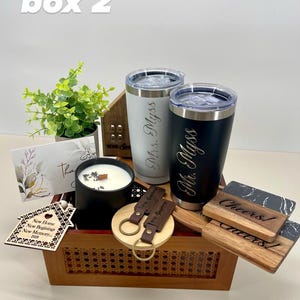 K&ouml;nnte beinhalten: Eine Holzschenkbox gef&uuml;llt mit personalisierten Artikeln f&uuml;r ein Einweihungsgeschenk. Die Box enth&auml;lt zwei gravierte Becher, eine Kerze, ein Holzhaus-Ornament, einen Schl&uuml;sselanh&auml;nger und zwei Untersetzer. Die Becher sind mit "Mr. Myss" und "Mrs. Myss" graviert. Die Untersetzer sind mit "Cheers!" und "Cheers!" graviert.