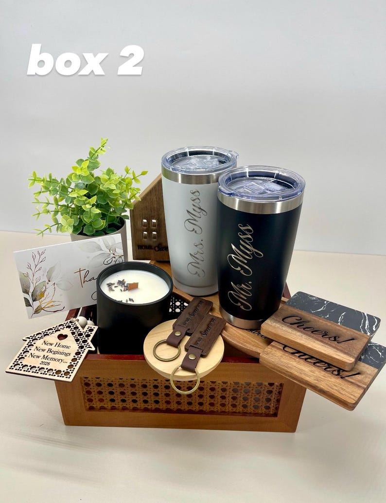 K&ouml;nnte beinhalten: Eine Holzschenkbox gef&uuml;llt mit personalisierten Artikeln f&uuml;r ein Einweihungsgeschenk. Die Box enth&auml;lt zwei gravierte Becher, eine Kerze, ein Holzhaus-Ornament, einen Schl&uuml;sselanh&auml;nger und zwei Untersetzer. Die Becher sind mit "Mr. Myss" und "Mrs. Myss" graviert. Die Untersetzer sind mit "Cheers!" und "Cheers!" graviert.