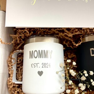 Puede incluir: Una taza blanca con la palabra "MOMMY" y "EST. 2024" impresas, junto con un s&iacute;mbolo de coraz&oacute;n. La taza est&aacute; en una caja de regalo con una tarjeta que dice "&iexcl;Felicidades!". La taza tiene una tapa y un asa plateadas.