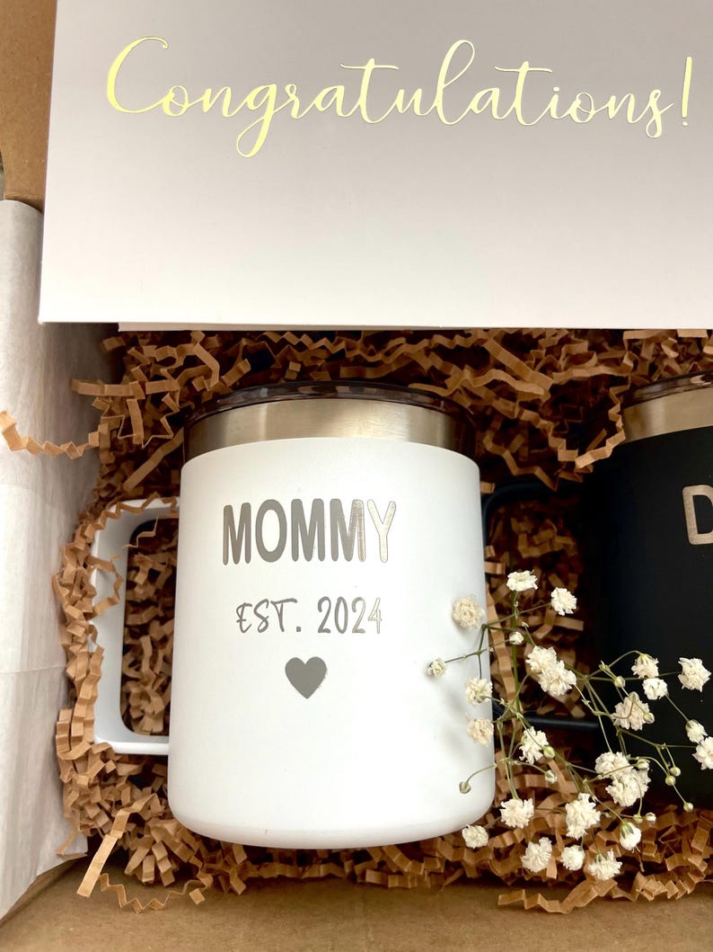 Puede incluir: Una taza blanca con la palabra "MOMMY" y "EST. 2024" impresas, junto con un s&iacute;mbolo de coraz&oacute;n. La taza est&aacute; en una caja de regalo con una tarjeta que dice "&iexcl;Felicidades!". La taza tiene una tapa y un asa plateadas.