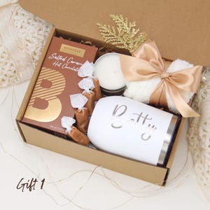 Puede incluir: Una caja de regalo llena con un vaso blanco con "Betty" en escritura plateada, chocolate caliente con caramelo salado, una vela, caramelos envueltos y un art&iacute;culo blanco esponjoso atado con una cinta dorada.