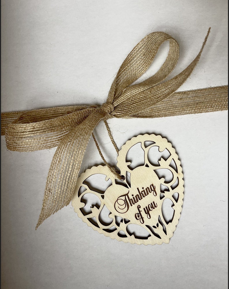 May include: A wooden heart-shaped tag with a cutout design and the text "Thinking of you" in a cursive font. The tag is tied to a brown burlap ribbon.