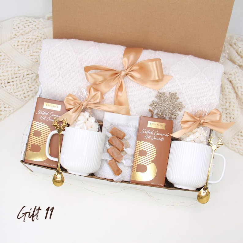 Puede incluir: Una caja de regalo con una manta blanca, dos tazas blancas con cucharas doradas, chocolate caliente con caramelo salado, malvaviscos y caramelos envueltos. La caja est&aacute; atada con una cinta dorada y tiene un adorno de copo de nieve.