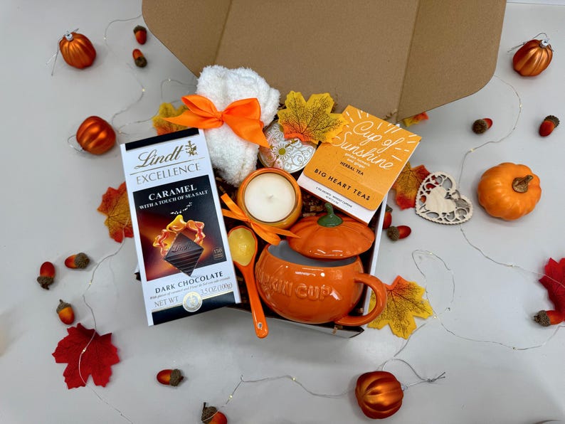 May include: An autumn-themed gift box filled with a pumpkin-shaped mug, candle, tea, chocolate bar, and a small white towel with an orange bow. Decorative pumpkins and leaves surround the box, creating a festive display.