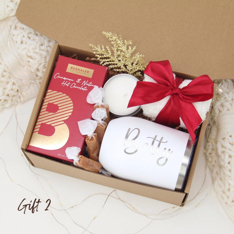 Puede incluir: Una caja de regalo que contiene un vaso blanco con "Betty" en escritura plateada, una caja roja de chocolate caliente, una vela peque&ntilde;a, caramelos envueltos, un lazo rojo y una ramita dorada. La caja es marr&oacute;n.