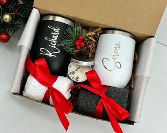 May include: A gift box filled with personalized items, including two stainless steel tumblers with the names "Richard" and "Serena" engraved on them, a small candle, a white towel, and gray socks. The box is lined with brown paper and has a red ribbon tied around it.