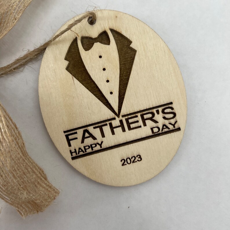 May include: A round wooden ornament with a laser-engraved tuxedo design and the text "Happy Father's Day 2023".