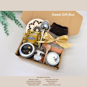 May include: A gift box with a variety of items, including a wooden coaster, Ghirardelli chocolates, honey, a wooden dipper, a handmade candle, cozy socks, natural black tea, and a tea infuser. The box is tied with a gold ribbon.