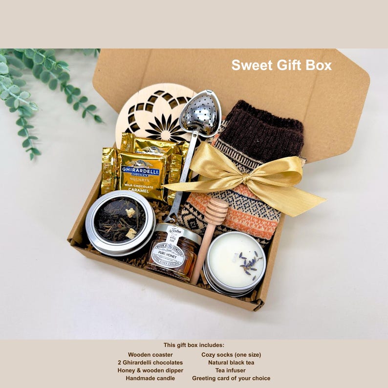 May include: A gift box with a variety of items, including a wooden coaster, Ghirardelli chocolates, honey, a wooden dipper, a handmade candle, cozy socks, natural black tea, and a tea infuser. The box is tied with a gold ribbon.