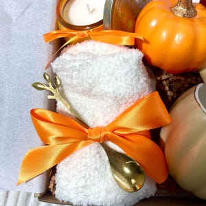 May include: A gift basket with a white towel, a gold spoon, a candle, a pumpkin spice tea bag, and a small orange pumpkin.
