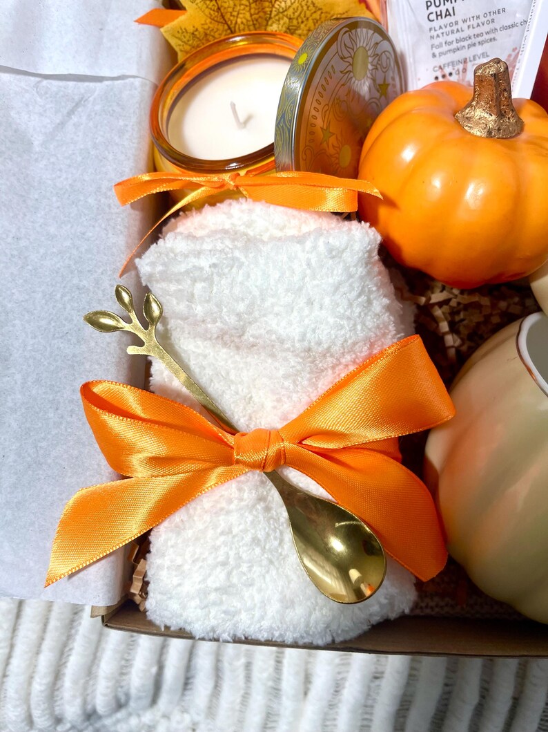 May include: A gift basket with a white towel, a gold spoon, a candle, a pumpkin spice tea bag, and a small orange pumpkin.