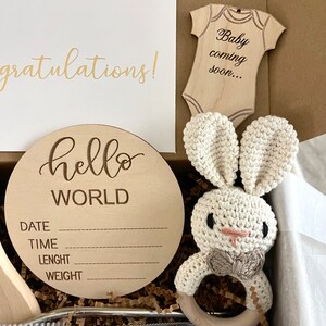 May include: A wooden baby announcement sign with the text "hello world" and spaces for the date, time, length, and weight. The sign is in a gift box with a crocheted bunny rattle, a wooden baby onesie cutout with the text "Baby coming soon...", and a card that says "Congratulations!"