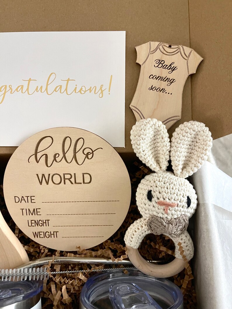 May include: A wooden baby announcement sign with the text "hello world" and spaces for the date, time, length, and weight. The sign is in a gift box with a crocheted bunny rattle, a wooden baby onesie cutout with the text "Baby coming soon...", and a card that says "Congratulations!"