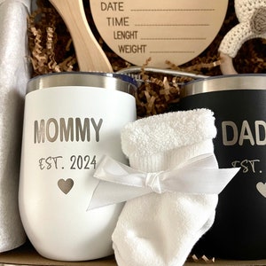 May include: A gift basket for new parents with two stainless steel tumblers, one white with "Mommy Est. 2024" and the other black with "Dad Est.", a white baby sock, a wooden sign that says "Hello World" with spaces to fill in the date, time, length, and weight of the baby, a wooden brush, and a crocheted bunny rattle.