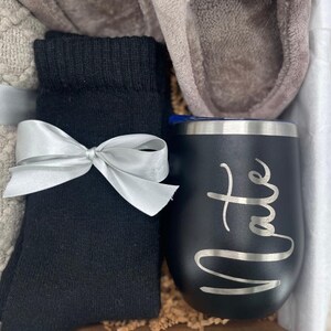 May include: A gift basket with a black and silver insulated tumbler with the name "Nate" engraved on it, a pair of black socks, a pair of gray slippers, and a white and gray blanket.