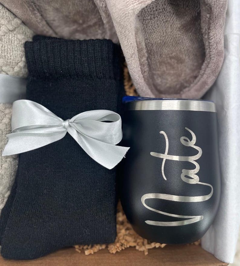 May include: A gift basket with a black and silver insulated tumbler with the name "Nate" engraved on it, a pair of black socks, a pair of gray slippers, and a white and gray blanket.