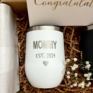Puede incluir: Un vaso aislado blanco con la palabra "MOMMY" y "EST. 2024" impresas, junto con un s&iacute;mbolo de coraz&oacute;n. El vaso est&aacute; en una caja de regalo con una tarjeta que dice "Congratulations" en letras doradas.