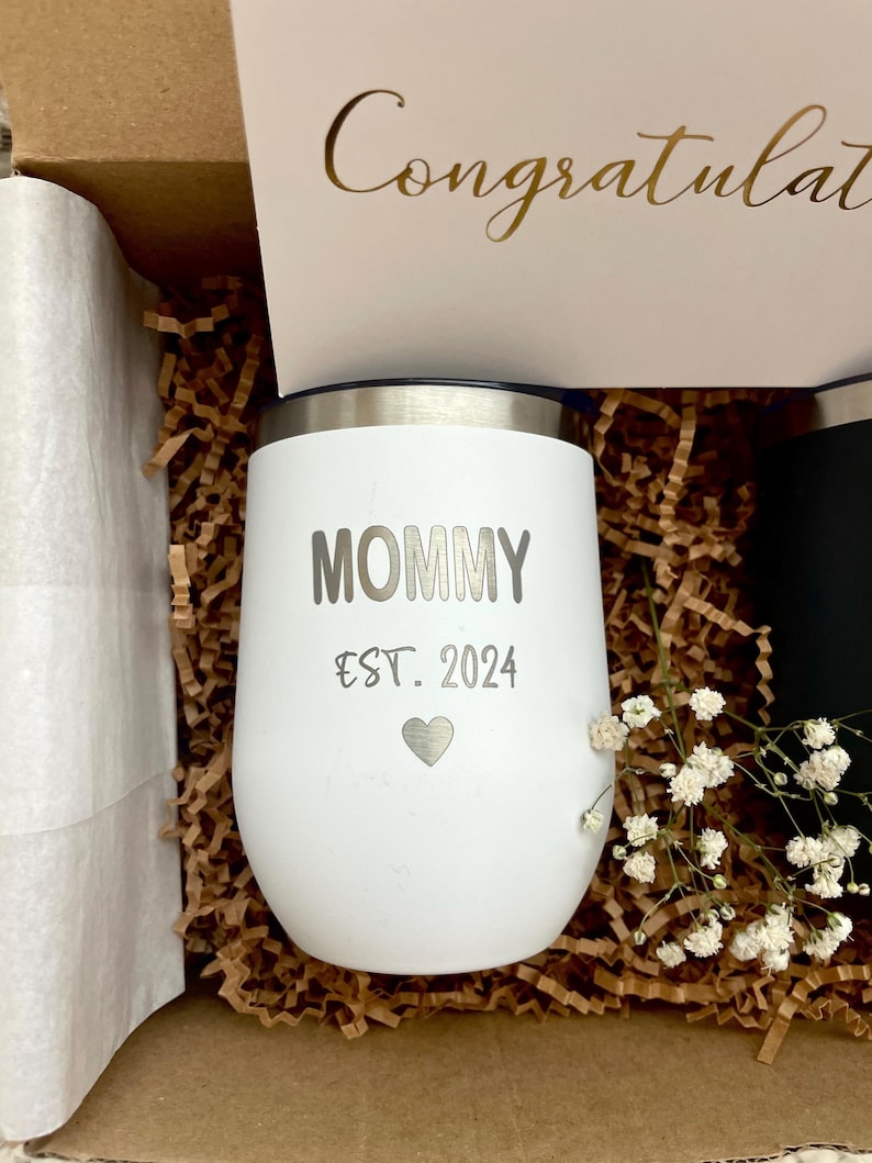 Puede incluir: Un vaso aislado blanco con la palabra "MOMMY" y "EST. 2024" impresas, junto con un s&iacute;mbolo de coraz&oacute;n. El vaso est&aacute; en una caja de regalo con una tarjeta que dice "Congratulations" en letras doradas.