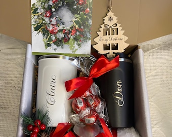 May include: A Christmas gift box with a card that says "Merry Christmas" and a wreath illustration. The box contains a white tumbler with "Claire" and a black tumbler with "Don", both with gold lettering. Also included are a wooden tree ornament, candies, and candles.