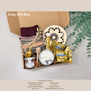 May include: A cozy gift box filled with a variety of items. The box includes a pair of cozy socks, a handmade candle, Ghirardelli chocolates, honey, a wooden dipper, a golden spoon, lip balm, and a wooden coaster. The box is labeled "Cozy Gift Box".