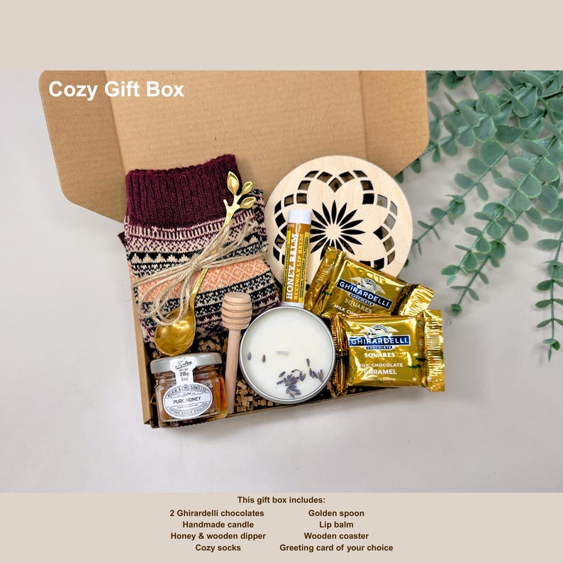 May include: A cozy gift box filled with a variety of items. The box includes a pair of cozy socks, a handmade candle, Ghirardelli chocolates, honey, a wooden dipper, a golden spoon, lip balm, and a wooden coaster. The box is labeled "Cozy Gift Box".