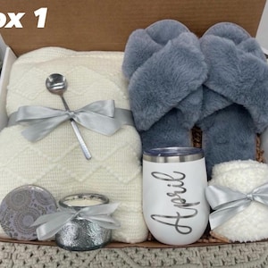 May include: A gift box labeled "box 1" containing a cream-colored blanket, gray slippers, a white tumbler with "April" in script, a candle, a spoon, and a small, fluffy item, all tied with silver ribbons.