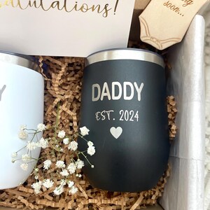Puede incluir: Caja de regalo con un vaso negro que dice "DADDY EST. 2024" con un coraz&oacute;n, y un vaso blanco. La caja tambi&eacute;n contiene una tarjeta que dice "&iexcl;Felicidades!" y un body de madera que dice "Baby coming soon..."