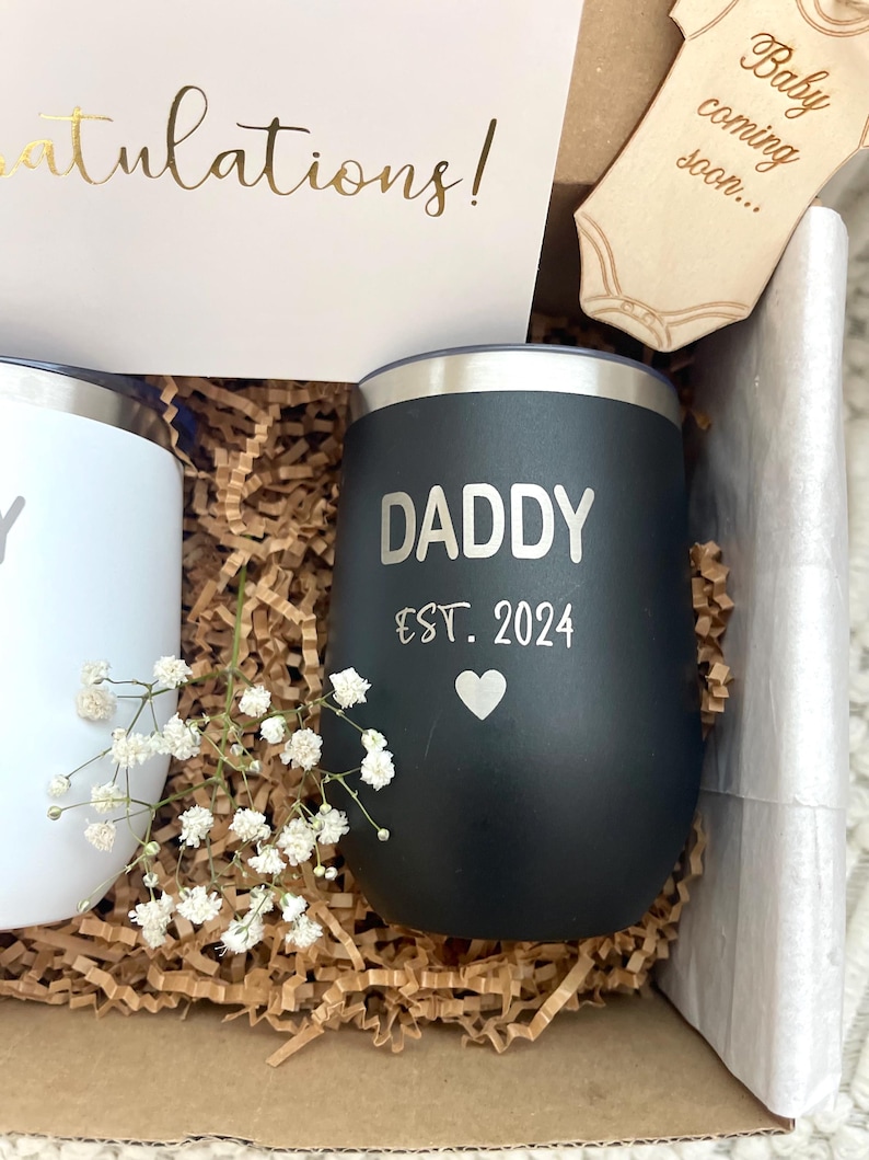 Puede incluir: Caja de regalo con un vaso negro que dice "DADDY EST. 2024" con un coraz&oacute;n, y un vaso blanco. La caja tambi&eacute;n contiene una tarjeta que dice "&iexcl;Felicidades!" y un body de madera que dice "Baby coming soon..."