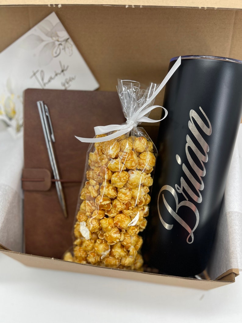 May include: A gift box filled with a brown leather journal, a silver pen, a bag of caramel popcorn, and a black stainless steel tumbler with the name "Brian" engraved on it.