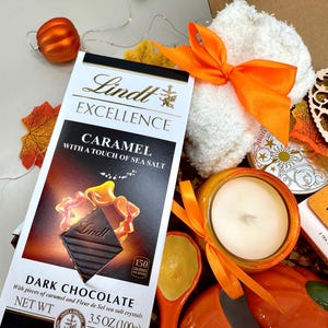 May include: A gift box with fall-themed items. Includes a Lindt Excellence dark chocolate bar with caramel and sea salt, a white washcloth tied with an orange ribbon, a candle, and a wooden heart with the text "Sending you sunshine."