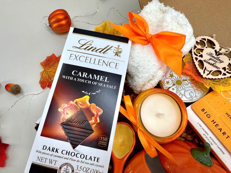 May include: A gift box with fall-themed items. Includes a Lindt Excellence dark chocolate bar with caramel and sea salt, a white washcloth tied with an orange ribbon, a candle, and a wooden heart with the text "Sending you sunshine."