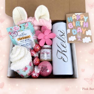 May include: A gift box containing a pink headband with ears, a floral face mask, a pink flower hair clip, a white tumbler with the name Kelsi, a Happy Birthday card, a jar of sugar scrub, a cherry blossom bath bomb, and four red wrapped candies.