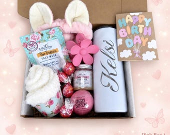 May include: A gift box containing a pink headband with ears, a floral face mask, a pink flower hair clip, a white tumbler with the name Kelsi, a Happy Birthday card, a jar of sugar scrub, a cherry blossom bath bomb, and four red wrapped candies.