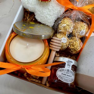 May include: A gift box with a candle, honey, chocolates, and a small towel. The candle is in an orange jar, and the honey is in a small jar with a label. The chocolates are wrapped in gold foil. The box is tied with an orange ribbon.