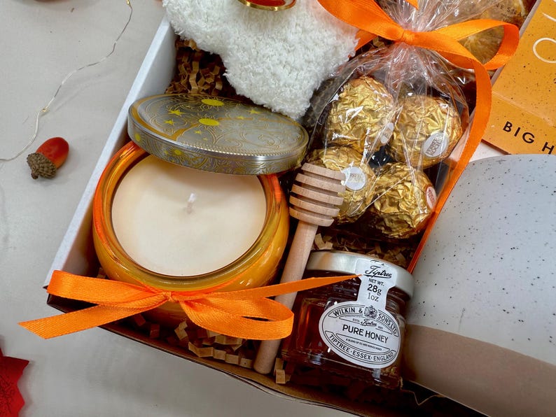 May include: A gift box with a candle, honey, chocolates, and a small towel. The candle is in an orange jar, and the honey is in a small jar with a label. The chocolates are wrapped in gold foil. The box is tied with an orange ribbon.