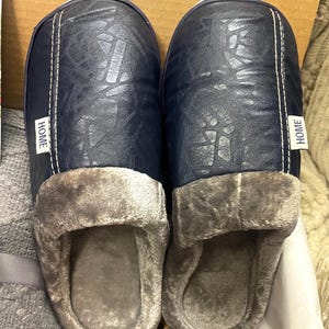 Pu&ograve; includere: Un paio di pantofole blu e grigie con tomaia in similpelle e fodera soffice. Le pantofole hanno un'etichetta "HOME" su ogni lato.