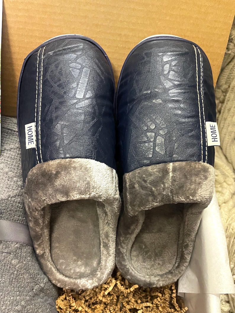 Pu&ograve; includere: Un paio di pantofole blu e grigie con tomaia in similpelle e fodera soffice. Le pantofole hanno un'etichetta "HOME" su ogni lato.