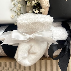 May include: A white baby sock with a white satin bow tied around it. The sock is sitting on top of a black and white striped ribbon. The sock is part of a baby gift basket.